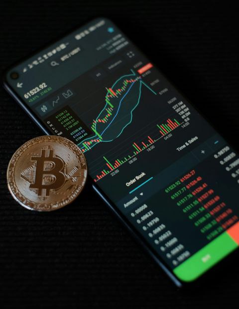 A close-up of a gold Bitcoin coin placed on a smartphone displaying a stock market chart.
